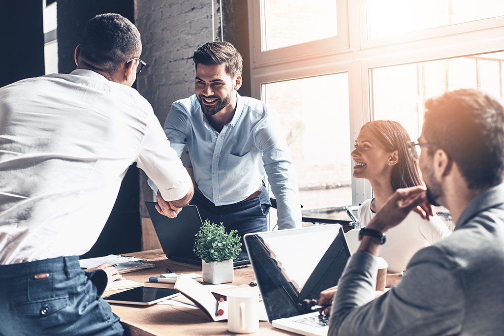 A business meeting with 4 people, smiling.  Two of them are shaking hands.