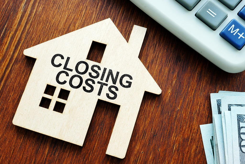 Closing costs written on a cardboard cutout of a home adjacent to a calculator and a stack of money.