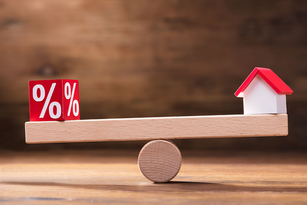 Interest rate percent sign on a balance beam with a home with the balance weighing slightly more on the interest rate percent sign side.