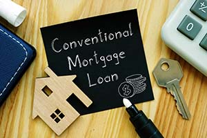 The words, "conventional loan mortgage" written on a note sitting on a desk next to a cardboard cutout of a house, a house key, a calculator, and a pen.