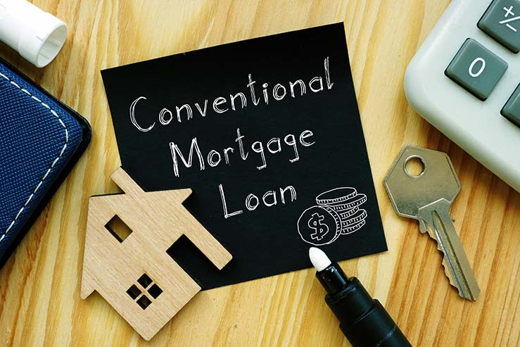 The words, "conventional loan mortgage" written on a note sitting on a desk next to a cardboard cutout of a house, a house key, a calculator, and a pen.